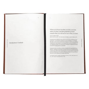 May include: An open brown leather-bound journal with a black ribbon bookmark. The text on the page reads "When you focus on what you lack, you lose what you have, and this gratitude journal ensures that you will never lose what you have. What is Gratitude? Gratitude is a gift you can give yourself every single day."