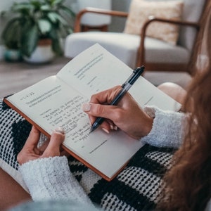 May include: A person writing in a journal with a black pen. The journal has a brown cover and white pages. The text in the journal reads "I'm grateful always for this moment to remember that form it takes".