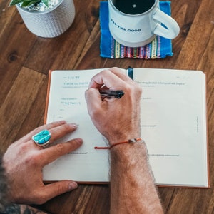 May include: A person's hand writing in a gratitude journal with a black pen. The journal has a tan cover and the pages are white. The text on the page reads "THANK YA THANK" and "The struggle ends when gratitude begins".