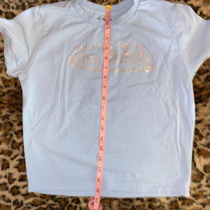 May include: A light blue short-sleeve t-shirt featuring the text Van Dutch in silver rhinestones across the chest. A pink measuring tape is placed vertically over the center of the shirt, showing a length of approximately 20 inches from the collar.