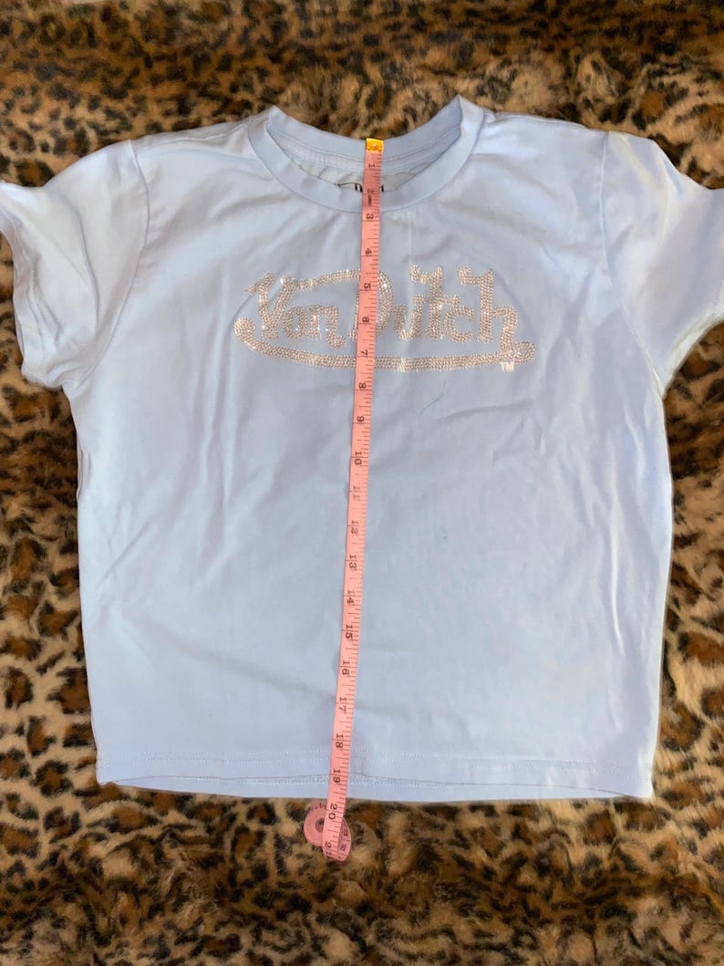 May include: A light blue short-sleeve t-shirt featuring the text Van Dutch in silver rhinestones across the chest. A pink measuring tape is placed vertically over the center of the shirt, showing a length of approximately 20 inches from the collar.