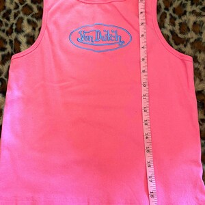 May include: A bright pink ribbed tank top featuring a blue embroidered Von Dutch logo in an oval frame on the chest. A black brand label is at the neckline. A pink measuring tape is placed vertically, showing a length of approximately 20 inches.
