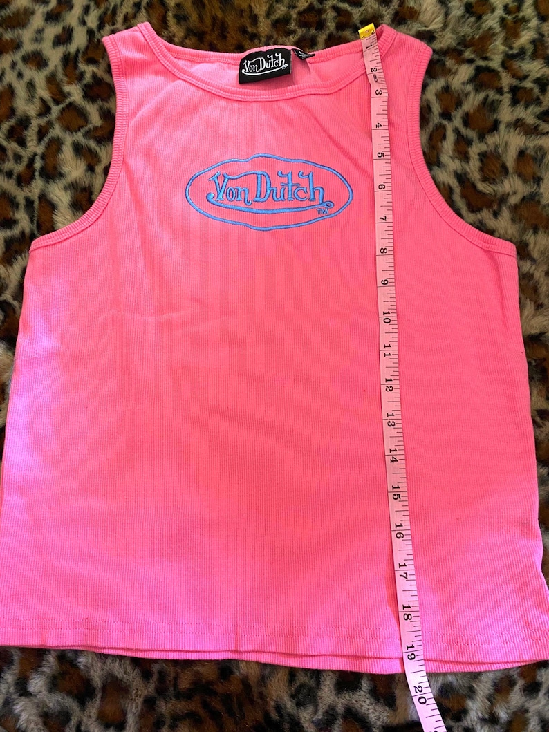 May include: A bright pink ribbed tank top featuring a blue embroidered Von Dutch logo in an oval frame on the chest. A black brand label is at the neckline. A pink measuring tape is placed vertically, showing a length of approximately 20 inches.
