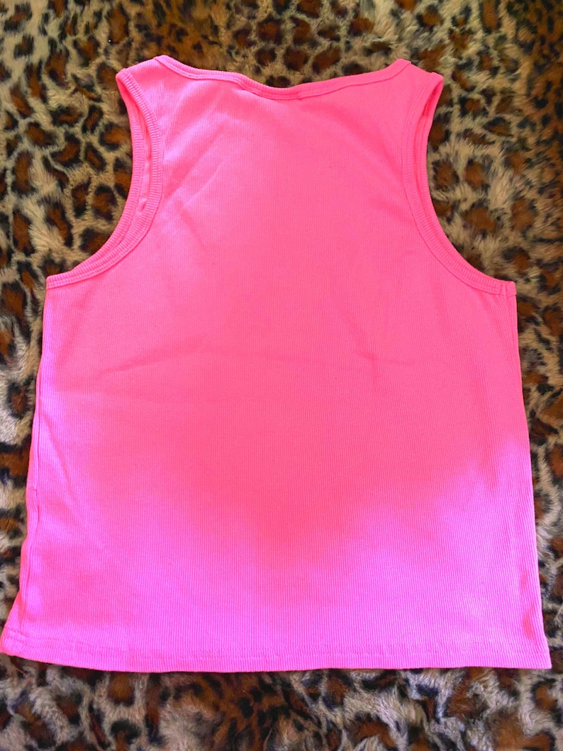 May include: A bright neon pink sleeveless tank top featuring a ribbed texture throughout the fabric. The garment has a classic scoop neckline and wide shoulder straps, with a straight hemline at the bottom, providing a simple and clean design.