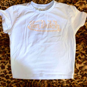 May include: A light blue short-sleeved t-shirt featuring the text Von Dutch written across the chest in a sparkling, light-colored rhinestone embellishment. The shirt has a crew neckline and a standard fit, with the brand logo clearly visible.