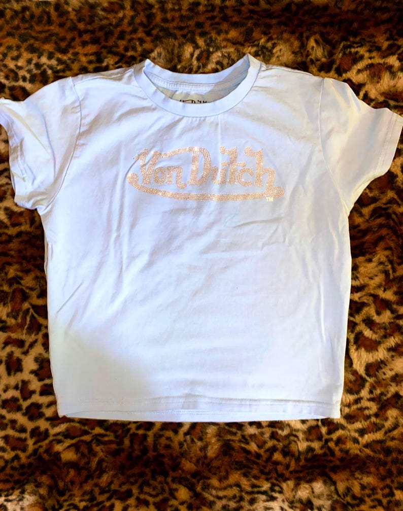 May include: A light blue short-sleeved t-shirt featuring the text Von Dutch written across the chest in a sparkling, light-colored rhinestone embellishment. The shirt has a crew neckline and a standard fit, with the brand logo clearly visible.