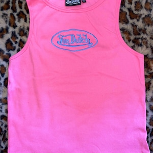 May include: A bright pink sleeveless tank top featuring a ribbed texture and a round neckline. The center of the chest displays an embroidered blue oval logo with the text Von Dutch. A black interior neck label also reads Von Dutch with size L.