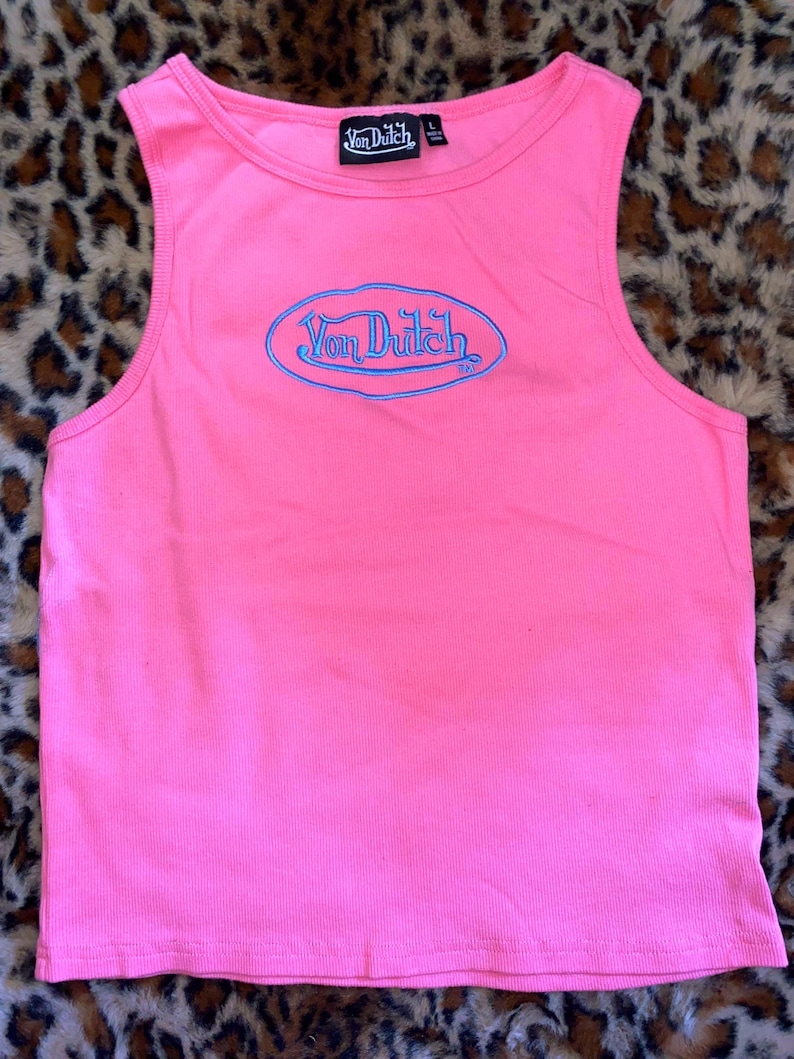 May include: A bright pink sleeveless tank top featuring a ribbed texture and a round neckline. The center of the chest displays an embroidered blue oval logo with the text Von Dutch. A black interior neck label also reads Von Dutch with size L.