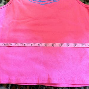May include: A bright pink ribbed tank top featuring a light blue graphic design near the neckline. A white measuring tape is placed horizontally across the midsection, indicating a width of approximately 16 inches from edge to edge.