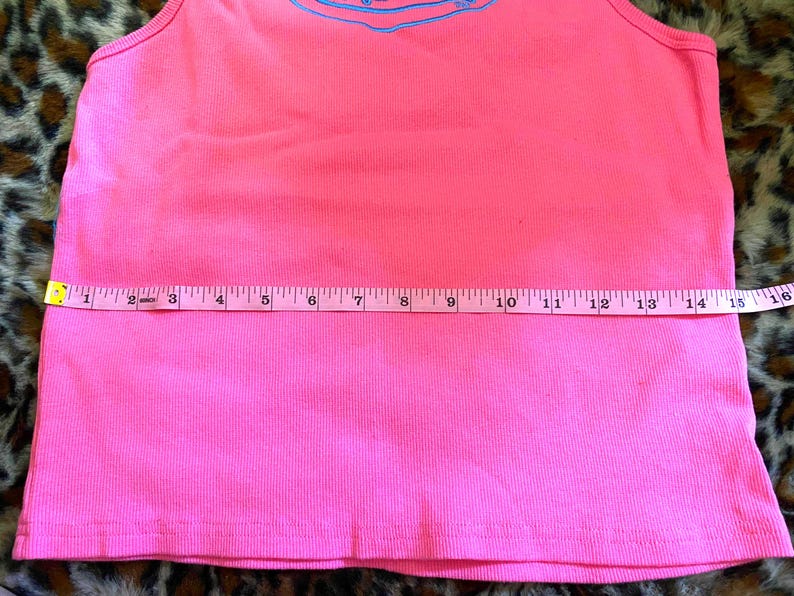 May include: A bright pink ribbed tank top featuring a light blue graphic design near the neckline. A white measuring tape is placed horizontally across the midsection, indicating a width of approximately 16 inches from edge to edge.