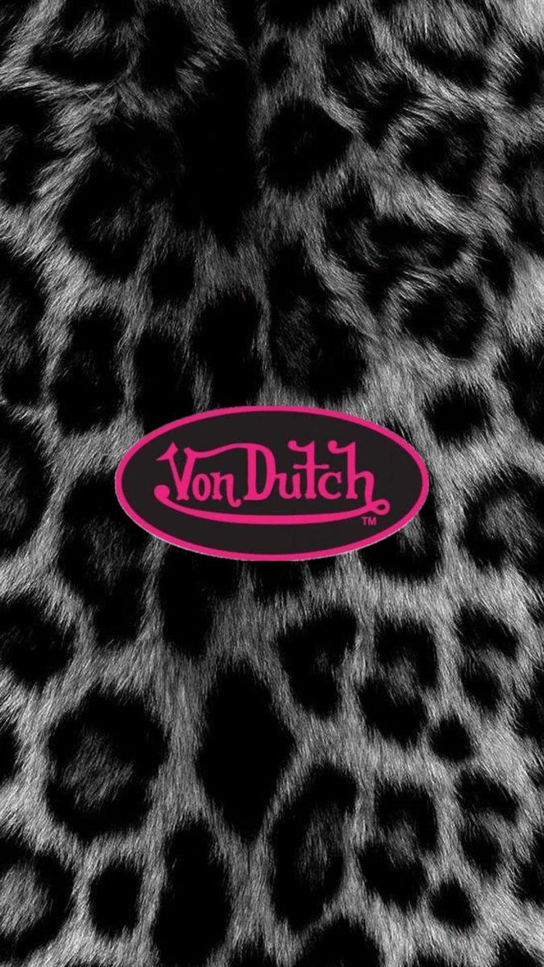 May include: A black and white leopard print pattern featuring a central horizontal oval emblem with a black background and a bright pink border. The text Von Dutch is written in a stylized pink script font inside the oval, followed by a small TM symbol.