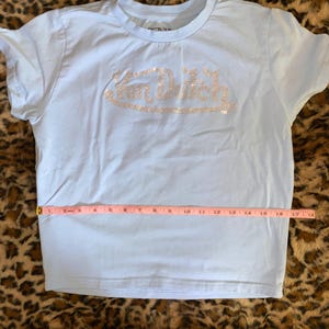 May include: A light blue short-sleeve t-shirt featuring the text Von Dutch in silver rhinestones across the chest. A pink measuring tape is placed horizontally across the midsection, showing a width measurement of approximately 18 inches.