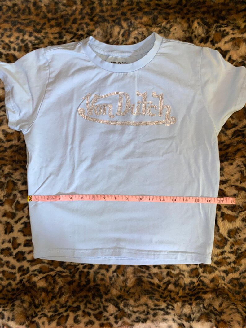 May include: A light blue short-sleeve t-shirt featuring the text Von Dutch in silver rhinestones across the chest. A pink measuring tape is placed horizontally across the midsection, showing a width measurement of approximately 18 inches.