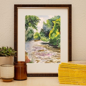 May include: A framed watercolor painting of a river flowing through a forest. The painting is in a brown frame with a white mat. The river is a light blue color and the trees are a dark green color.