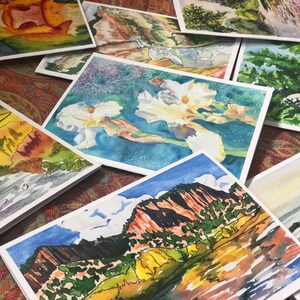 May include: A collection of watercolor paintings on cards, featuring landscapes, flowers, and abstract designs. The cards are white with a slight texture and are arranged in a pile.