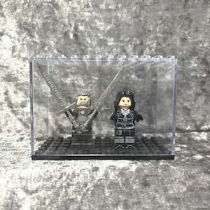 May include: Two miniature figures of a man and a woman, both wearing dark clothing and holding swords, are displayed in a clear plastic display case with a black base.