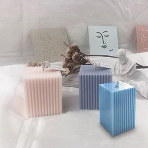 May include: Three square pillar candles with a ribbed texture. The candles are pink, blue and grey.