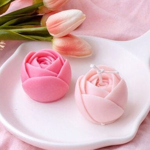 May include: Two pink rose-shaped candles on a white ceramic plate. The candles are made of wax and have a delicate, feminine design.