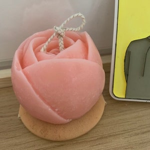 May include: A pink rose-shaped candle with a white string tied around the top. The candle is sitting on a small, round, light brown base.