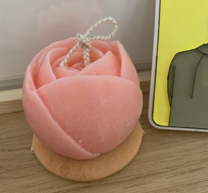 May include: A pink rose-shaped candle with a white string tied around the top. The candle is sitting on a small, round, light brown base.