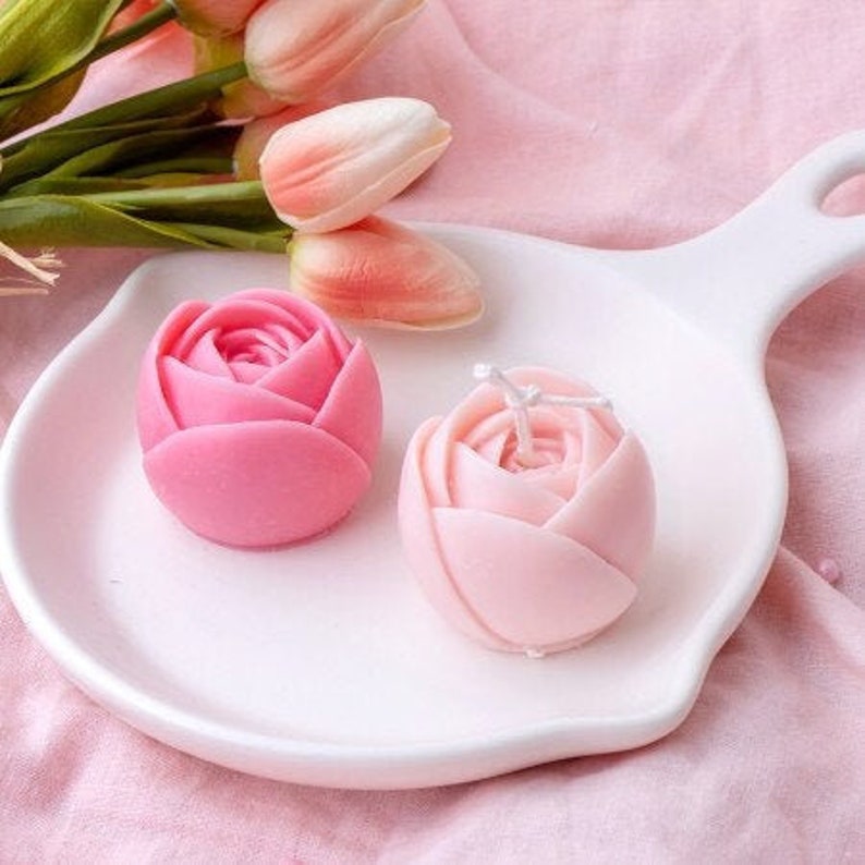 May include: Two pink rose-shaped candles on a white ceramic plate. The candles are made of wax and have a delicate, feminine design.