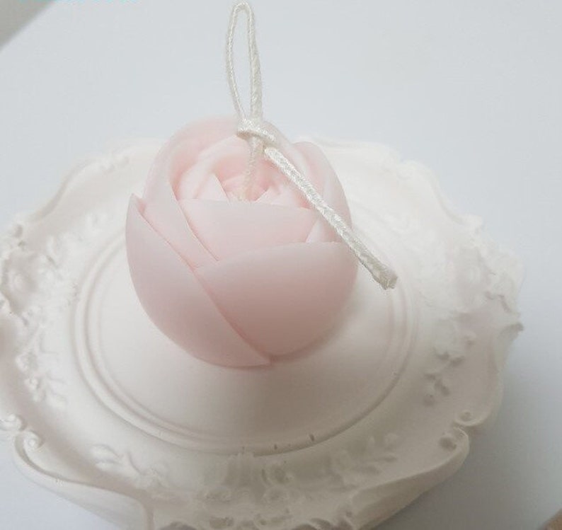 May include: A delicate pink rose-shaped candle with a white wick and a white string attached to the top.