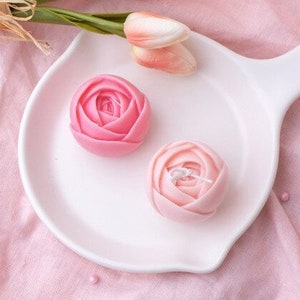May include: Two pink rose-shaped candles on a white plate. The candles are made of wax and have a realistic rose design.