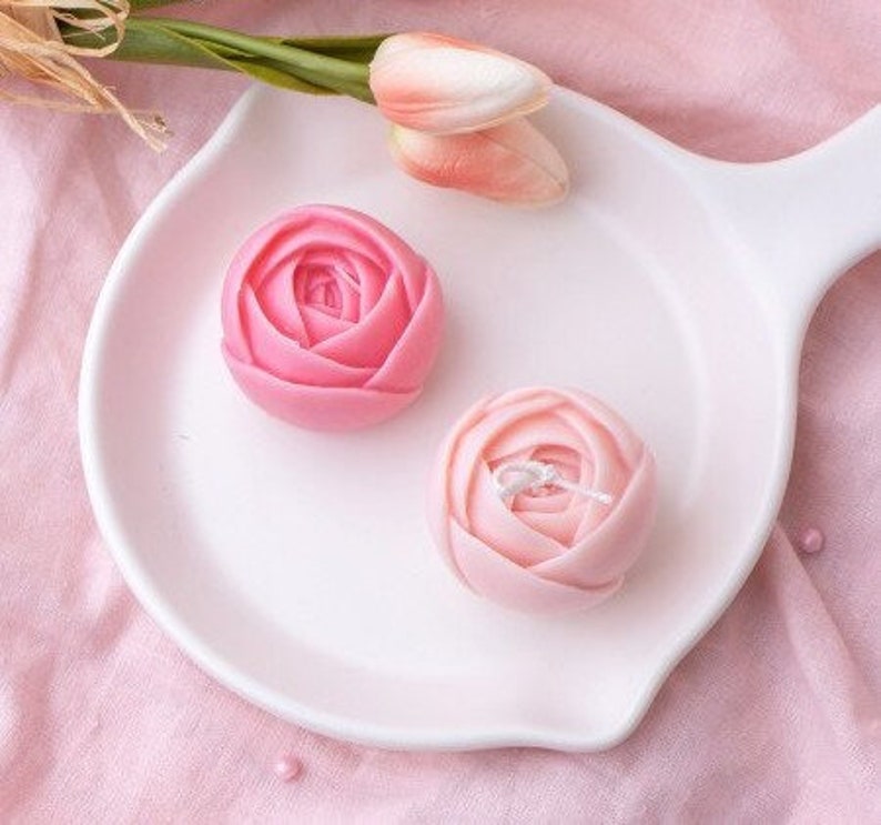 May include: Two pink rose-shaped candles on a white plate. The candles are made of wax and have a realistic rose design.