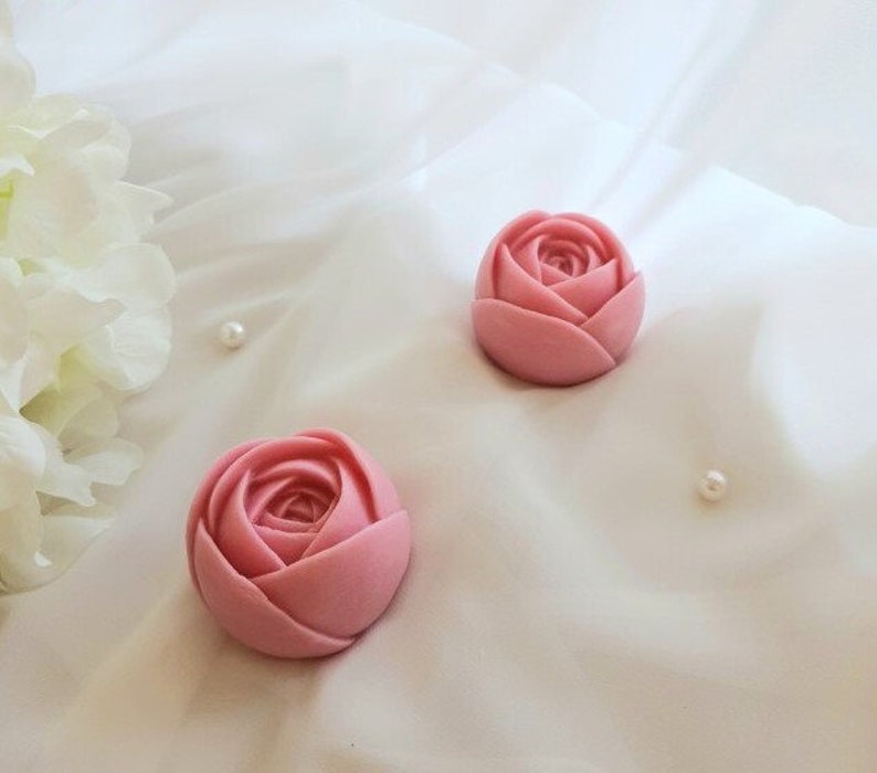 May include: Two pink rose-shaped soaps on a white background. The soaps are made to look like real roses.