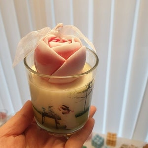 May include: A pink and white rose-shaped candle in a clear glass jar with a green and white floral design. The candle is topped with a white ribbon.