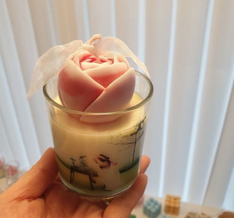 May include: A pink and white rose-shaped candle in a clear glass jar with a green and white floral design. The candle is topped with a white ribbon.