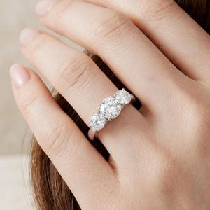 May include: A three-stone ring featuring a large round brilliant-cut centre diamond flanked by two smaller round diamonds, all set in a polished silver-toned metal band. The design showcases a classic prong setting for each stone.