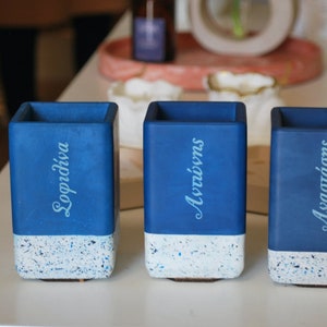 Puede incluir: Tres macetas de hormigón azul con fondo moteado blanco. Las macetas están etiquetadas con "Sopraviva", "Acrivivns" y "Asvaatadons" en una fuente azul claro.