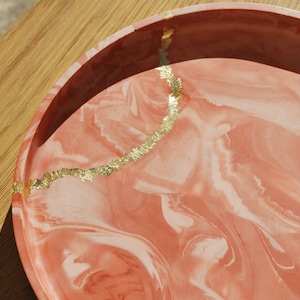 May include: A round, coral-colored tray with a marbled design and gold leaf accents.