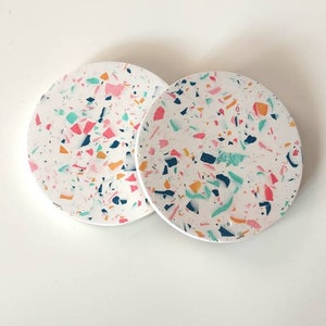 Könnte beinhalten: Zwei runde weiße Untersetzer mit einem bunten Terrazzo-Muster. Die Untersetzer haben einen Mix aus rosa, blauen, grünen und orangen Flecken.