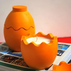 May include: Two orange egg-shaped candle holders with a white candle in one of them. The candle holders are sitting on a stack of books.