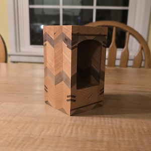 May include: A handcrafted wooden box with a rectangular shape and a decorative chevron pattern. The box features a cutout on one side and is made of light and dark brown wood. The box is approximately 6 inches tall.