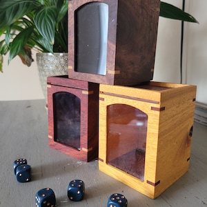 May include: Three wooden boxes with glass windows, stacked on top of each other. The boxes are made of different types of wood, including walnut, cherry, and maple. The boxes are empty and have a natural finish. There are five blue dice with gold dots on a gray surface in front of the boxes.