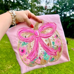 May include: A light pink t-shirt featuring a pumpkin design with a large pink bow. The pumpkin is decorated with floral patterns in shades of pink, teal, and gold glitter accents. The t-shirt is held up by a hand with red nail polish.