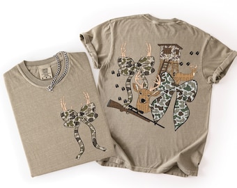 Puede incluir: Dos camisetas color beige con gráficos temáticos de caza. Una camiseta presenta un lazo de camuflaje con astas, la otra tiene un ciervo, un lazo, un rifle y una torre de caza. Ambas camisetas tienen una etiqueta Comfort Colors.