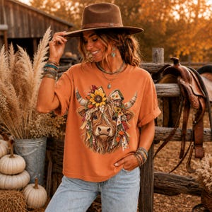 May include: A burnt orange short-sleeve t-shirt featuring a graphic print of a Highland cow adorned with a sunflower and colorful patterned ribbons. The shirt is paired with light blue denim jeans and a brown wide-brimmed felt hat.
