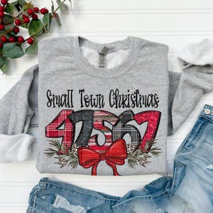 Könnte beinhalten: Grauer Pullover mit dem Text "Small Town Christmas" und den Zahlen 47567 in rot-schwarz-weißem Karomuster. Die Zahlen sind von einem Kranz aus Kiefernästen und einer roten Schleife umgeben.
