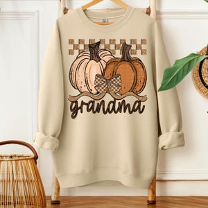 May include: Beige sweatshirt featuring a pumpkin graphic with a bow and the word "grandma" in a script font. The pumpkins are in shades of orange and peach, with a checkered pattern above them. The sweatshirt has long sleeves and a crew neck.