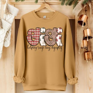 May include: Mustard-colored sweatshirt with the word "GIGI" in a plaid design with Christmas tree accents. The phrase "helping raise tiny disciples" is written in a script font below the word "GIGI".