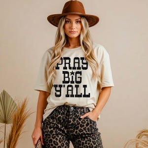 May include: A woman wearing a tan t-shirt with the text "PRAY BIG Y'ALL" in black block letters. She is also wearing a brown hat and leopard print jeans. The shirt is a casual, oversized style.