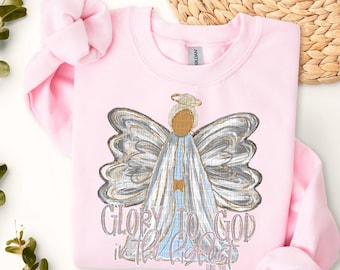 Könnte beinhalten: Rosafarbener Pullover mit einem Aufdruck eines Engels mit silbernen Flügeln und dem Text "Ehre sei Gott in der Höhe Lukas 2,14".