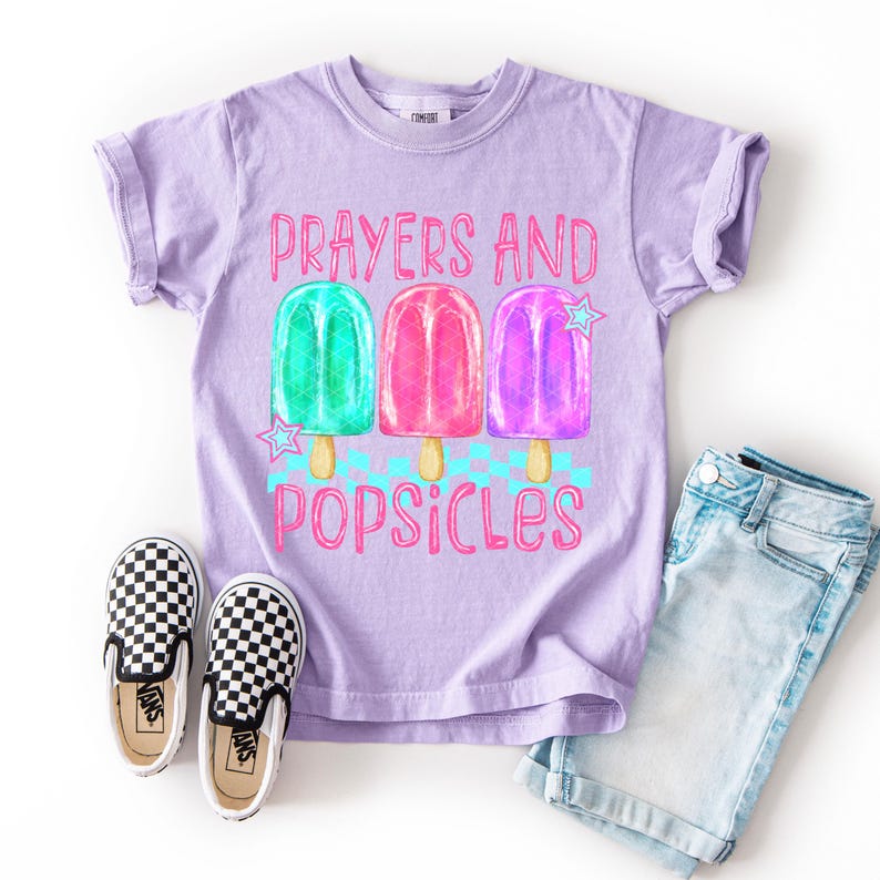 Może przedstawiać: Liliowa koszulka z r&oacute;żowym napisem "Prayers and Popsicles". Koszulka przedstawia ilustracje zielonych, r&oacute;żowych i fioletowych lod&oacute;w na patyku. Na zdjęciu znajduje się r&oacute;wnież para czarno-białych szachownicowych but&oacute;w wsuwanych i dżinsowych szort&oacute;w.