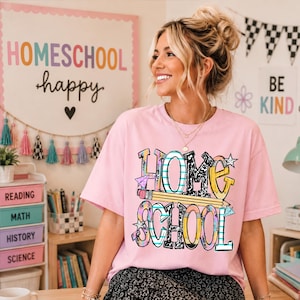 May include: A light pink short-sleeved t-shirt featuring a graphic print that reads "HOMESCHOOL" in large, stylized, multi-colored letters with patterns, including a yellow pencil illustration running horizontally through the center of the text.