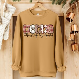 May include: Mustard yellow sweatshirt with the word "Mama" in a decorative font. The letters have plaid, tree, and patterned designs. Below the word "Mama" is the phrase "helping raise tiny disciples" in a script font.