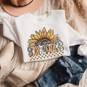 May include: White sweatshirt with a sunflower graphic and the text "find me among the SUNFLOWERS". The design features a large sunflower with yellow, orange, and brown petals. The sweatshirt is paired with blue jeans and beige boots.